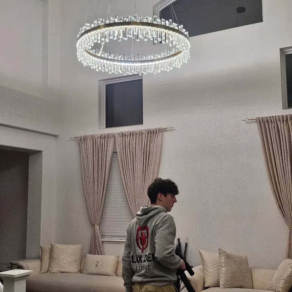 A person wearing a gray hoodie with an electrical-company logo stands in a high-ceiling living room beneath an illuminated circular crystal chandelier. Curtains and cream-colored seating surround them.