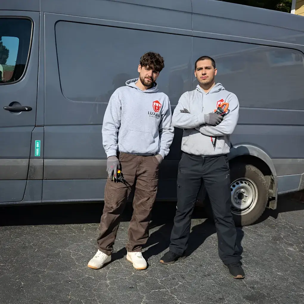 Two workers in matching logo hoodies and gloves pose with hand tools in front of a gray cargo van outdoors.