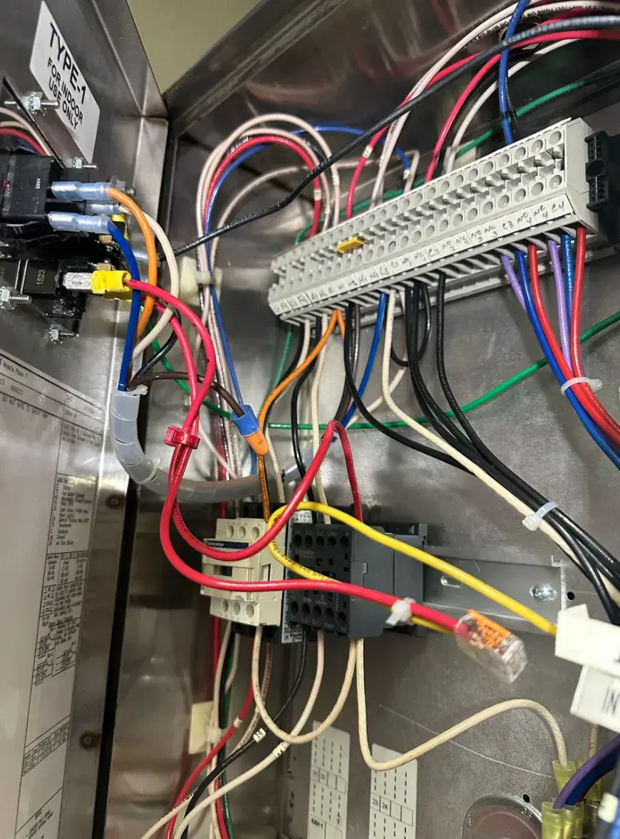 Close-up inside a metal electrical control cabinet, showing multicolored wiring, a labeled terminal strip, relays, and wire connectors.