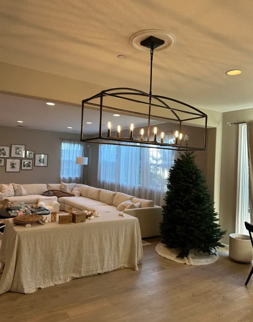 A lit black open-frame chandelier hangs above a covered dining table in a living area with recessed lights, a white sectional sofa, and a Christmas tree.