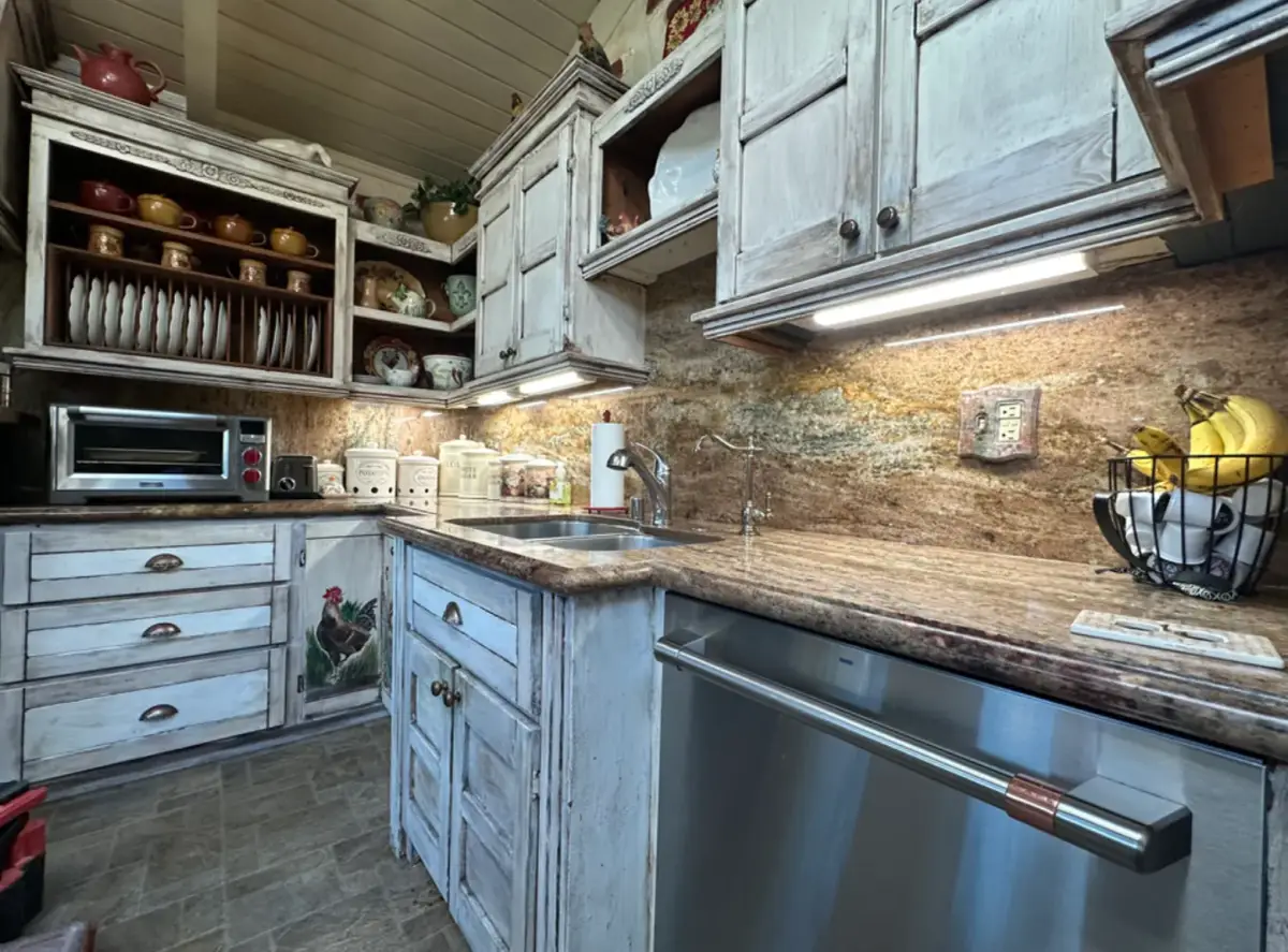 Kitchen with distressed white cabinets, patterned stone countertops and backsplash, under-cabinet lighting, a double sink, and a stainless steel dishwasher.