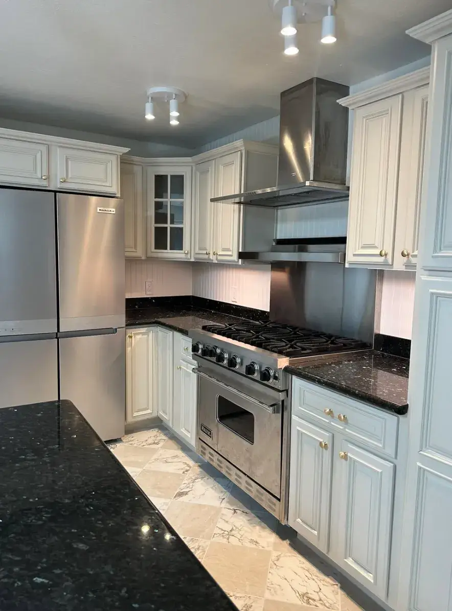 Kitchen with white paneled cabinets, dark stone countertops, a stainless steel refrigerator, gas range and large vent hood, and patterned tile flooring.