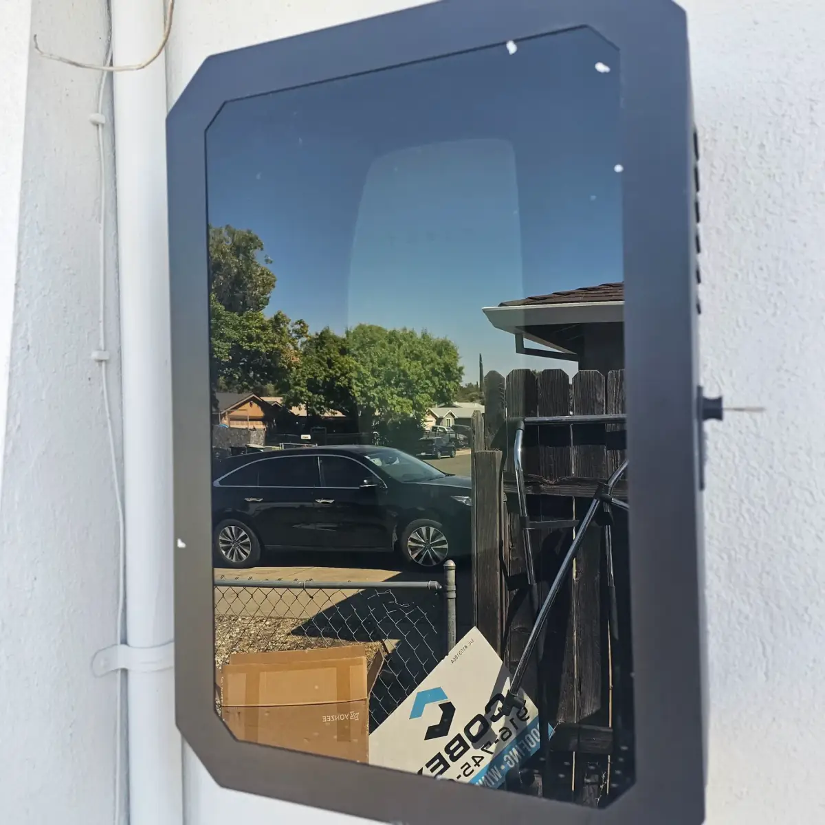 A dark, reflective-front enclosure mounted on a white exterior wall beside conduit. Its surface reflects a parked car, trees, fencing, and cardboard boxes.