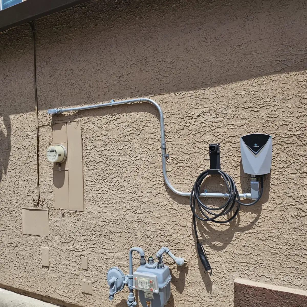 A wall-mounted Lectron electric vehicle charger with a coiled cable and hanging connector on a tan stucco exterior wall. Metal conduit runs beside an electrical meter and panel, with a gas meter below.