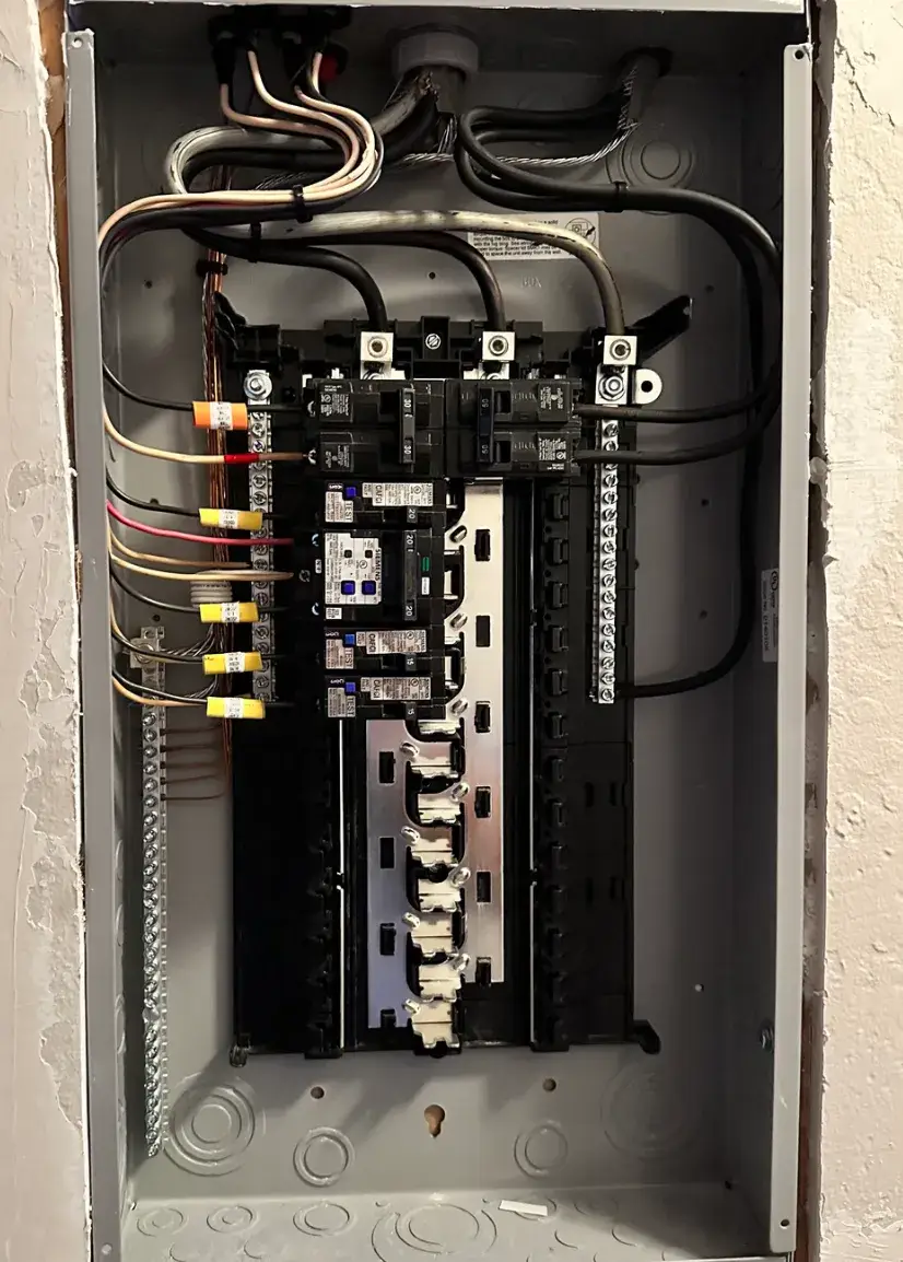 An open wall-mounted electrical panel shows circuit breakers, connected wiring, metal terminal bars, and unused breaker spaces.