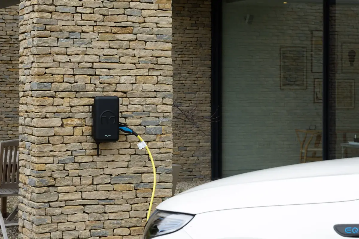 A black electric vehicle charger is mounted on an exterior stone wall, with a yellow cable beside the front of a white car.