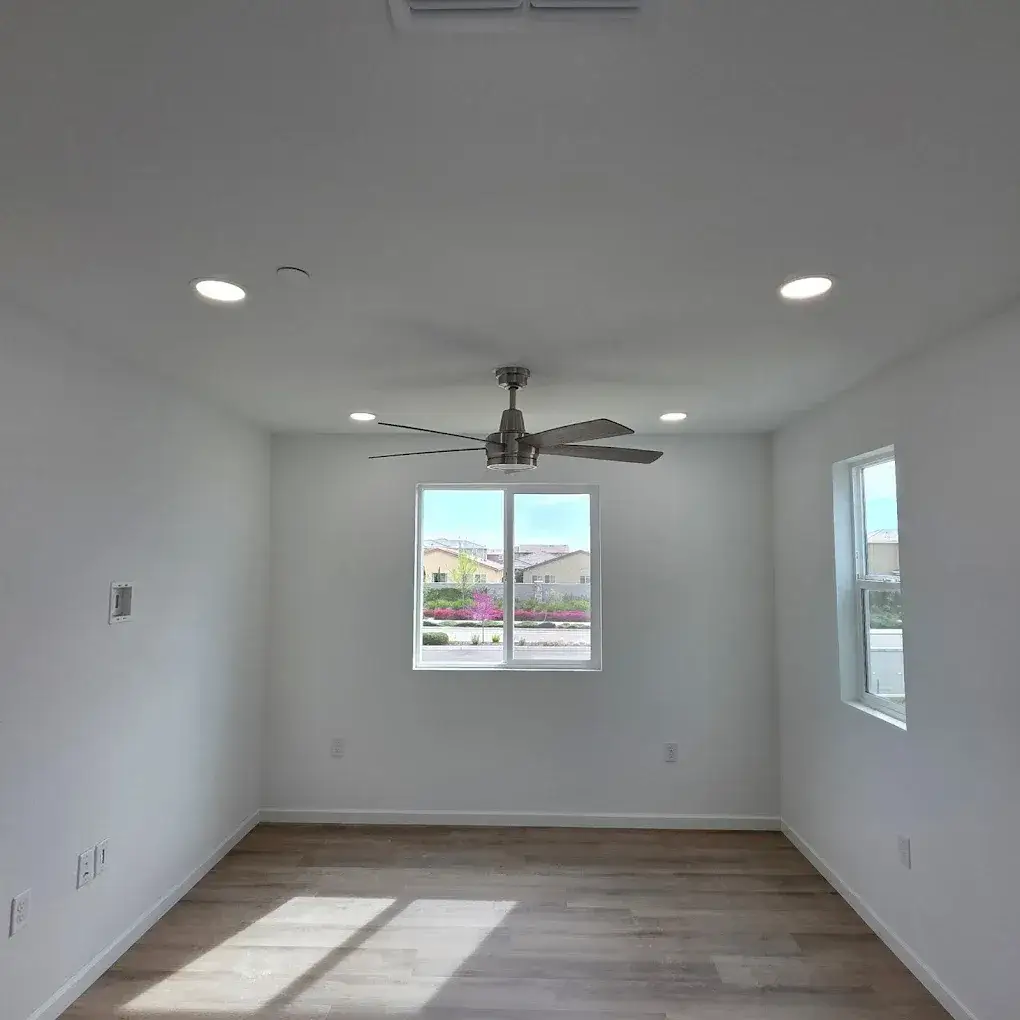 An empty room with white walls, wood-look flooring, two windows, illuminated recessed ceiling lights, and a ceiling fan.