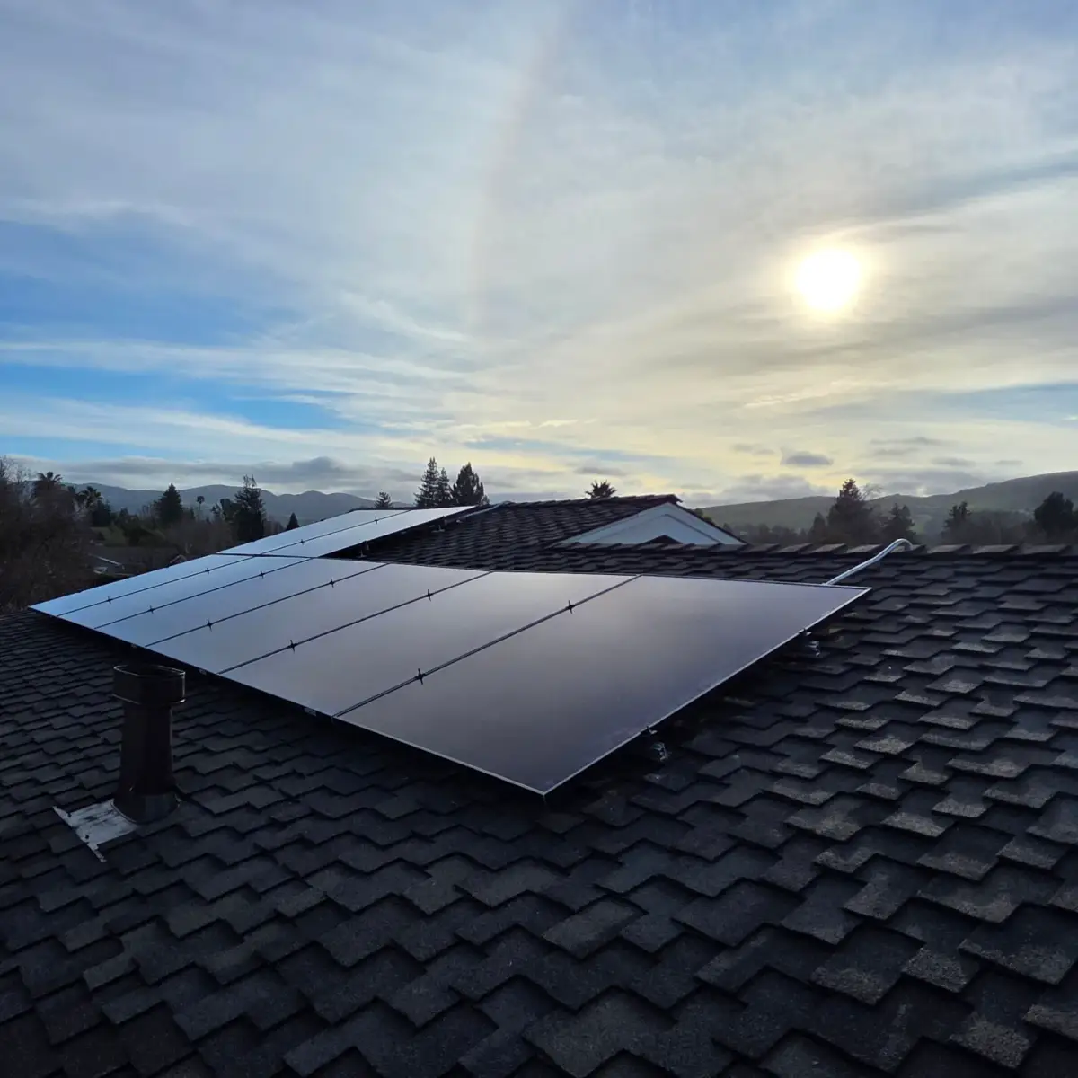 Solar panels mounted on a dark shingle roof, with a roof vent in front and trees, hills, and a bright cloudy sky behind.