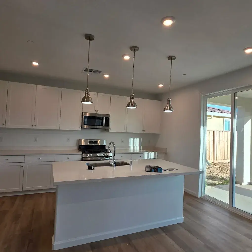 White kitchen with illuminated pendant and recessed lights, stainless steel appliances, an island sink, wood-look flooring, and sliding glass doors. Small tools and packages sit on the island.