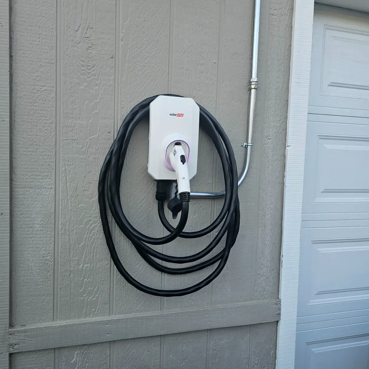 A white SolarEdge electric vehicle charger with a coiled black cable and docked connector is mounted on gray exterior siding beside a garage door. Metal conduit connects to the unit.