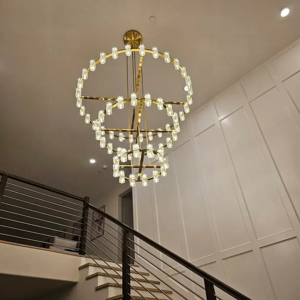 An illuminated gold-toned chandelier with crystal-like lights hangs above an indoor staircase with black metal railings and white paneled walls.