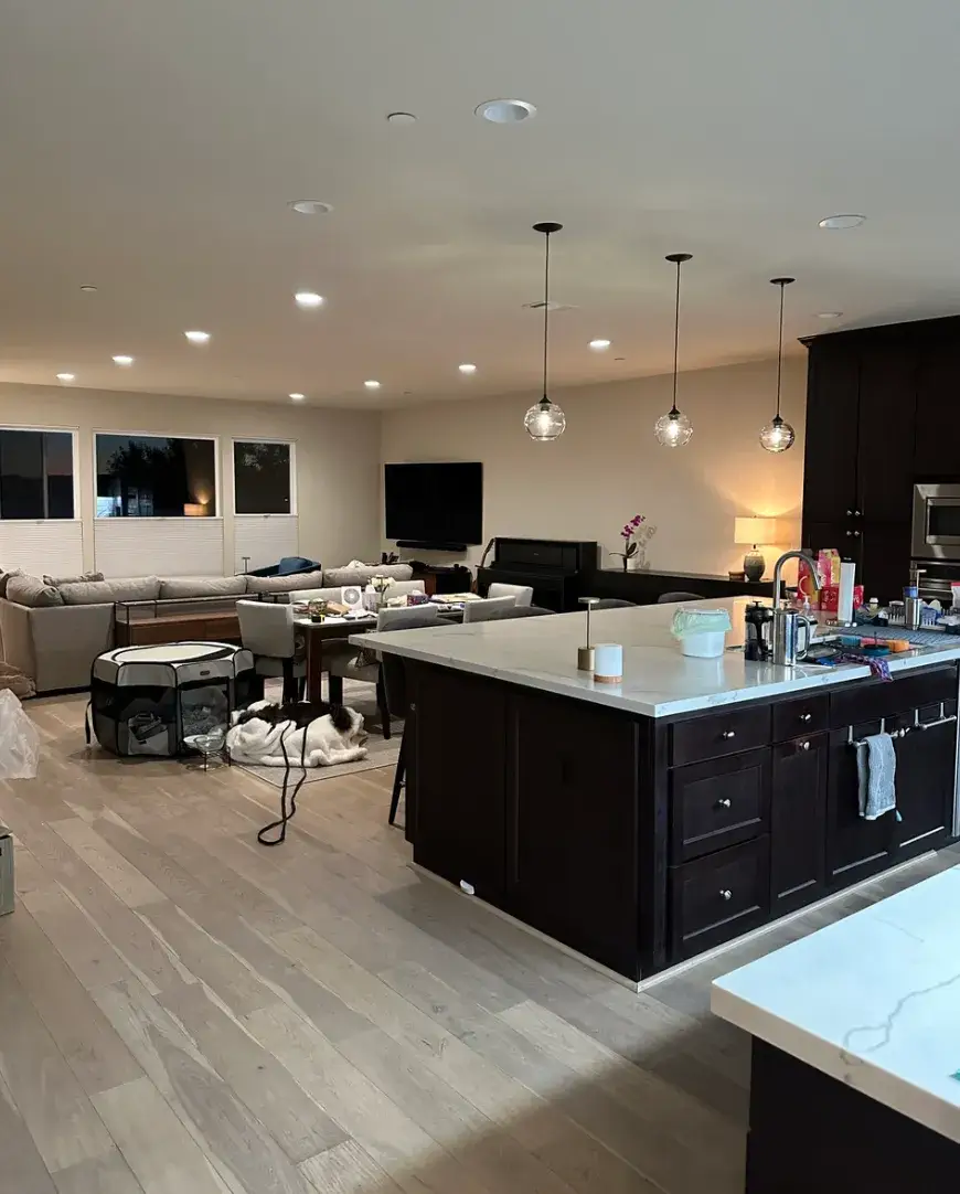 Open-plan kitchen and living room with dark cabinetry, a white island countertop, three glass pendant lights, recessed ceiling lights, and wood-look flooring. Furniture and household items fill the adjoining living space.
