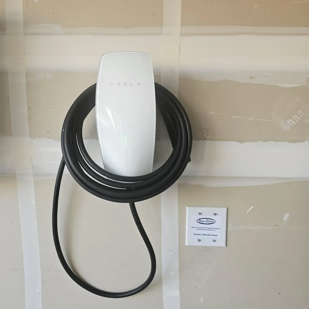 A white Tesla wall-mounted electric vehicle charger with a coiled black cable on unfinished drywall, beside a Rayco Electric label.