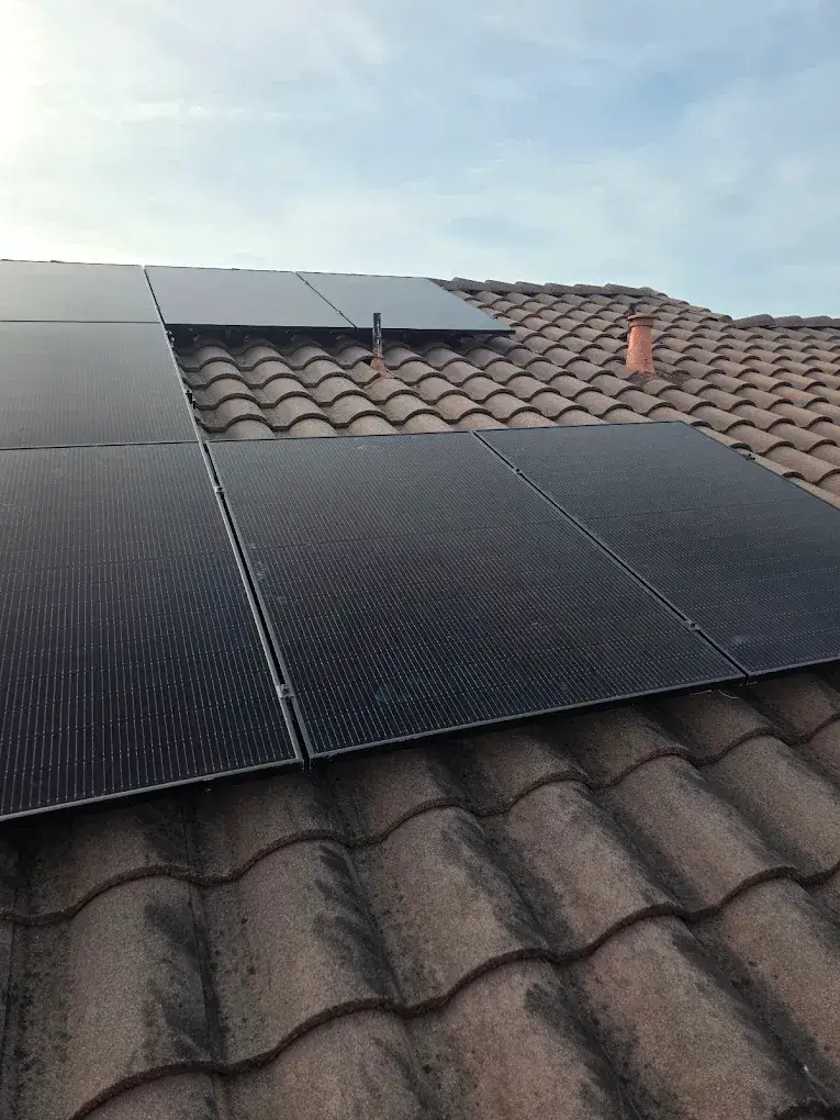 Black solar panels mounted on a sloped, curved-tile roof, with roof vents and cloudy sky visible.