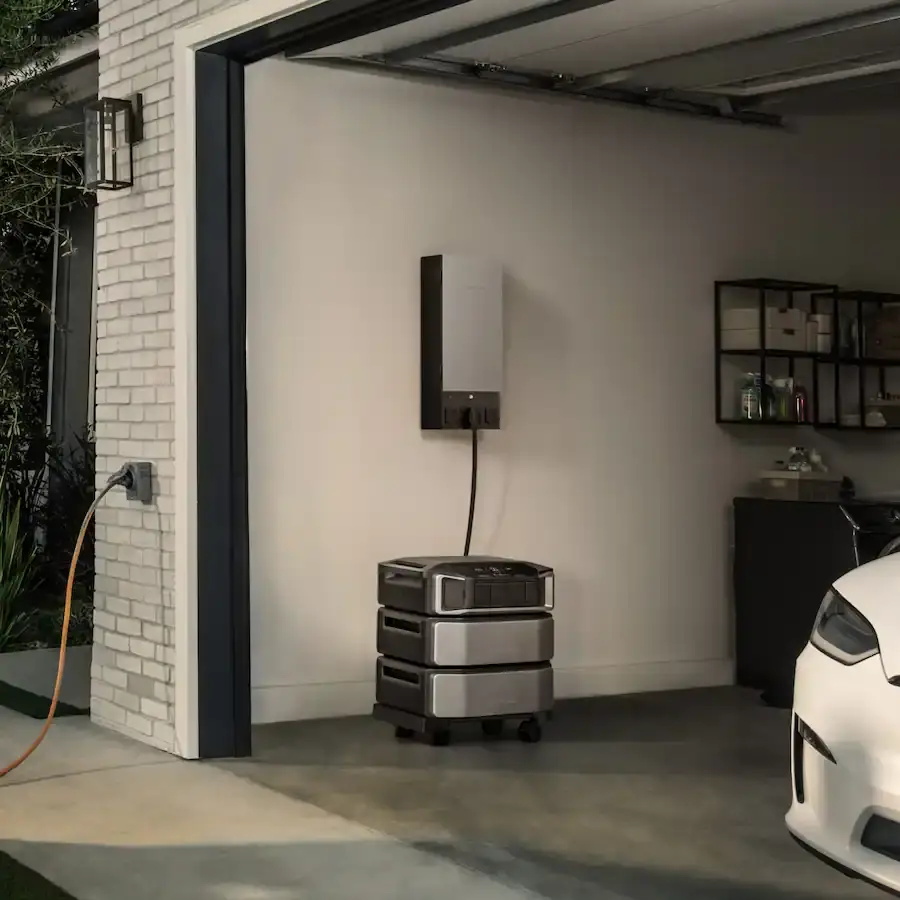 An open residential garage contains stacked portable power units connected to a wall-mounted device, shelving, and the front of a white car.