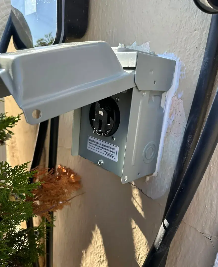 An outdoor electrical receptacle in a gray metal weatherproof box with its lid open, mounted on a stucco wall beside black cables and shrubs.