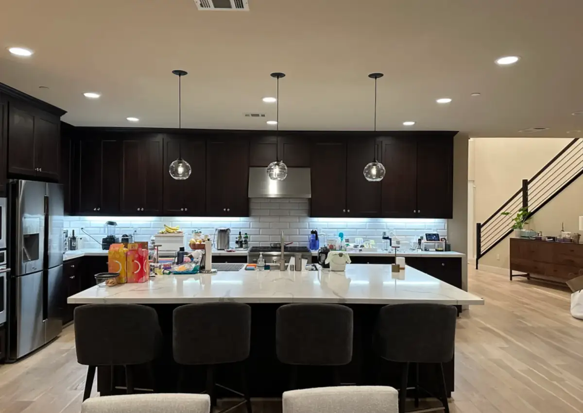 Kitchen with dark cabinets, a white island with four stools, stainless steel appliances, three glass pendant lights, and illuminated tile backsplash. Household items cover parts of the counters, and a staircase is visible at right.
