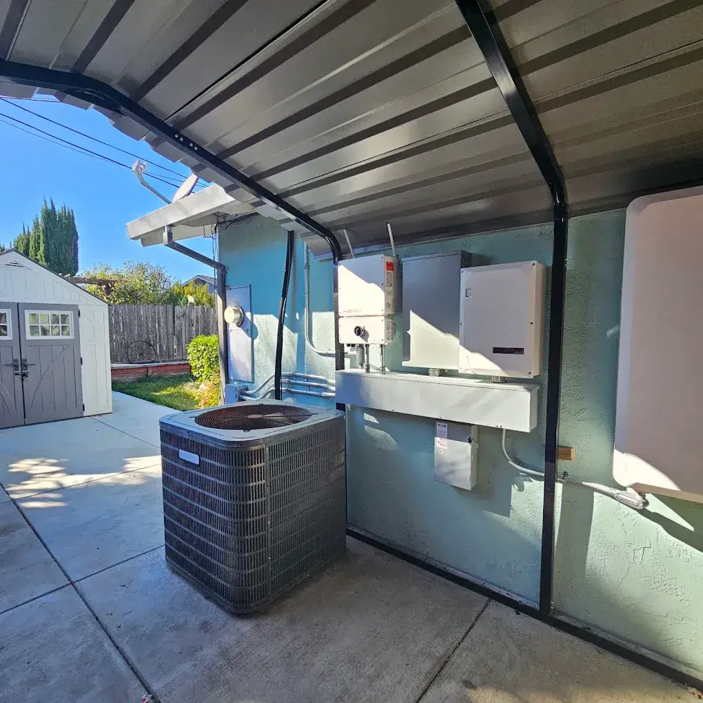 Outdoor air-conditioning condenser beside wall-mounted electrical equipment and conduit under a metal patio cover, with a small shed nearby.