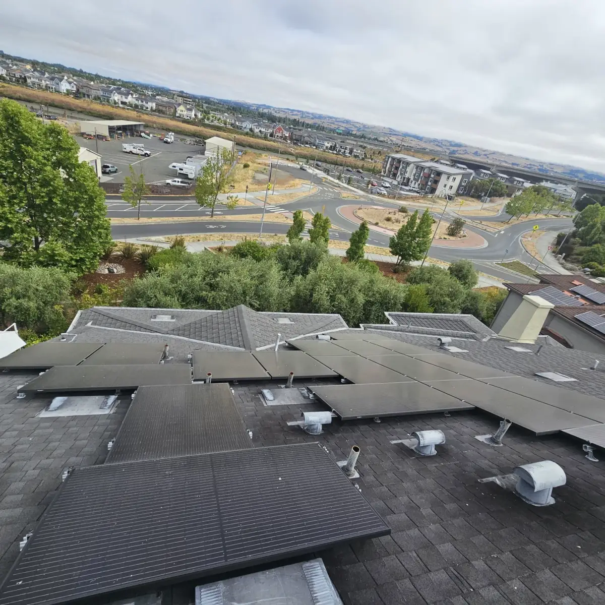 Solar panels mounted on a gray shingle roof among metal vents, overlooking trees, homes and a roundabout under cloudy skies.