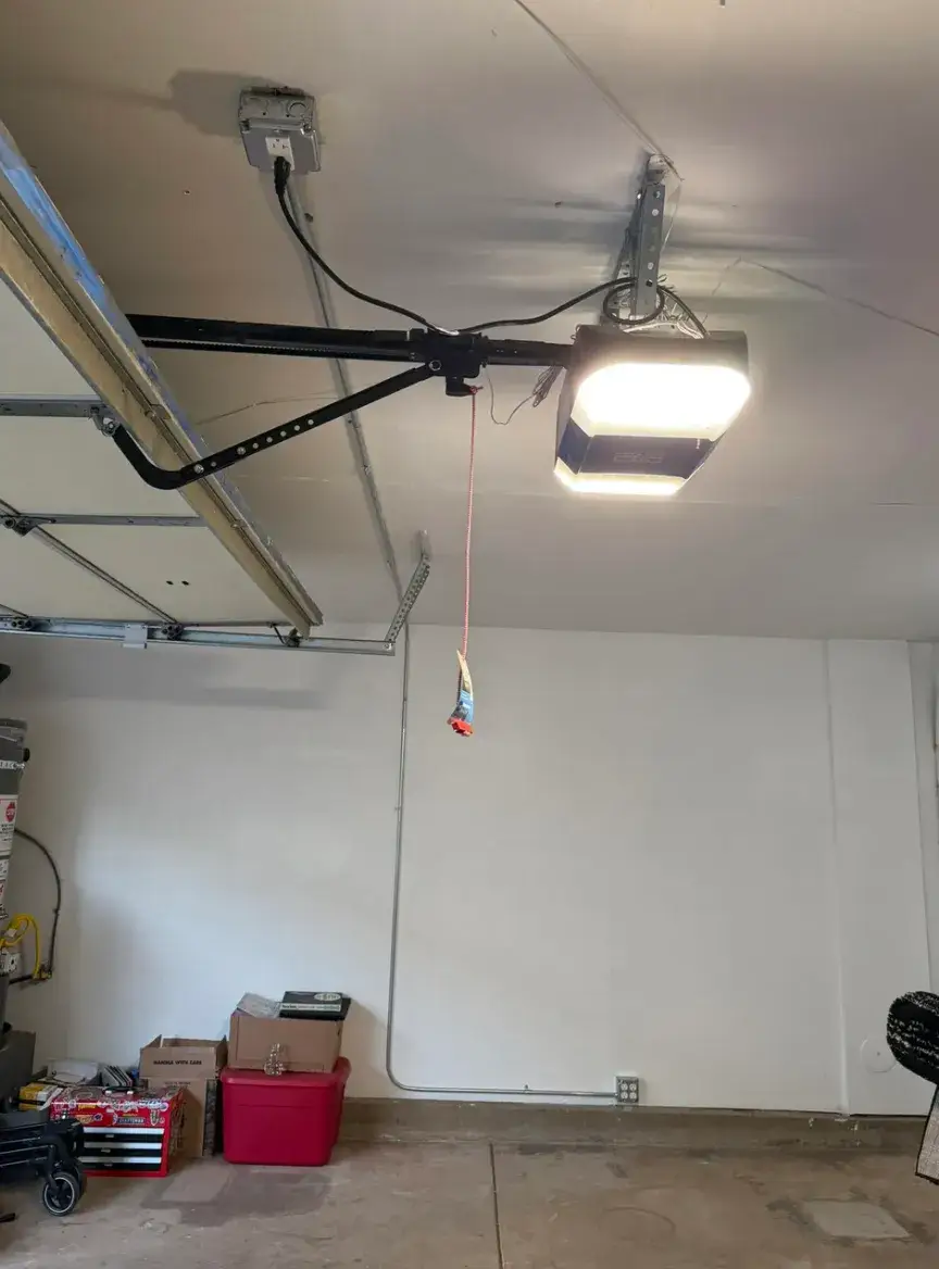 Garage interior with a ceiling-mounted garage door opener, illuminated lights, metal rails, and a hanging emergency release cord. Storage boxes and tools sit along the wall.