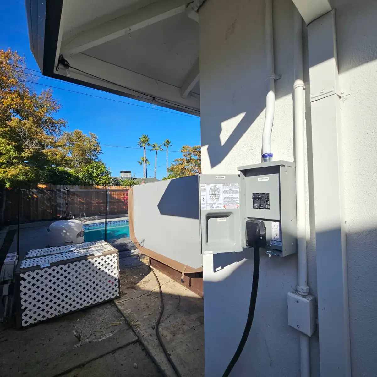 An outdoor electrical panel with its door open shows breakers, outlets and a plugged-in cable beside a backyard pool and covered hot tub.