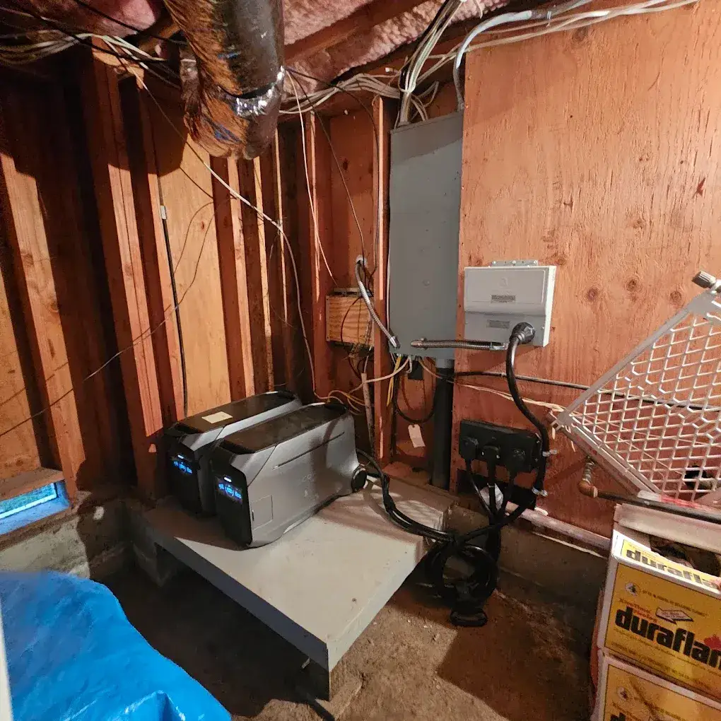 Two portable power units sit on a platform beside wall-mounted electrical equipment and connected cables in an unfinished basement with exposed framing and ductwork.