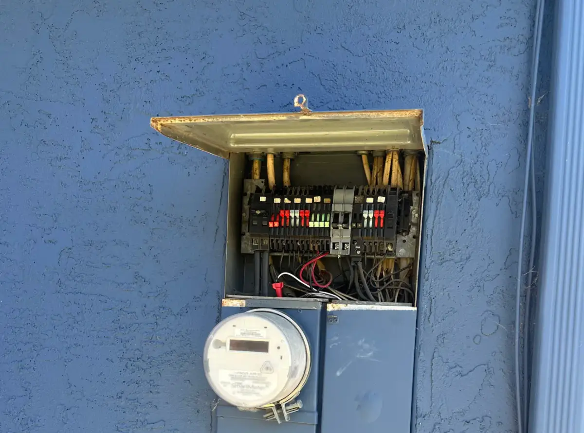 An open outdoor electrical panel exposes circuit breakers and wiring above a round electric meter on a blue stucco wall. Rust is visible along the raised metal lid.