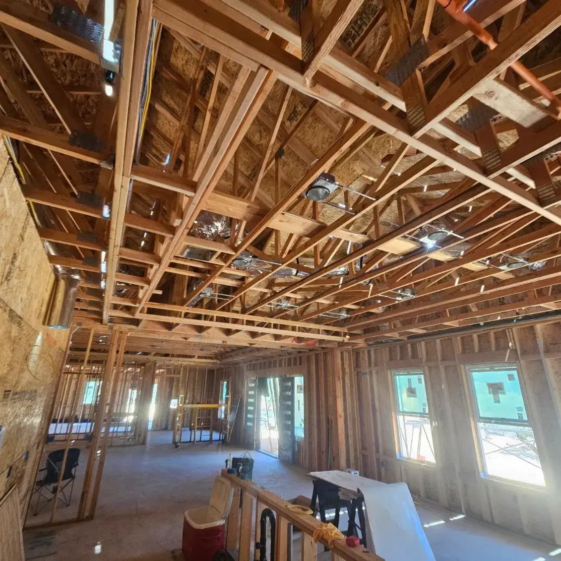 Unfinished building interior with exposed wood framing, ceiling ductwork, recessed light housings, windows, and construction supplies. No people are visible.