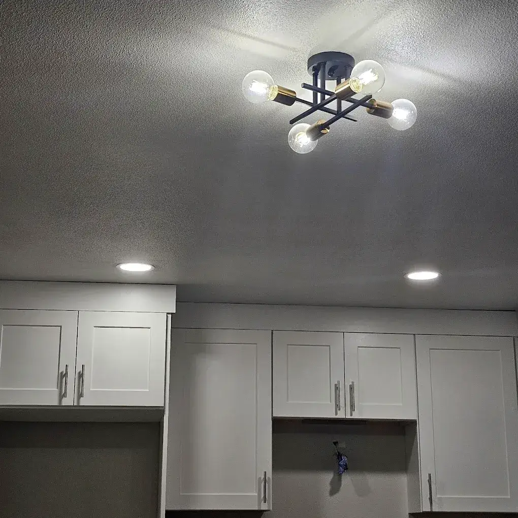 An illuminated black-and-brass ceiling fixture and two recessed lights above white kitchen cabinets, with an exposed wire visible below one cabinet.