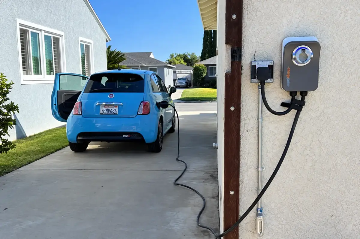 A wall-mounted electric vehicle charger connects by a long cable to a blue car parked in a residential driveway with its driver-side door open.