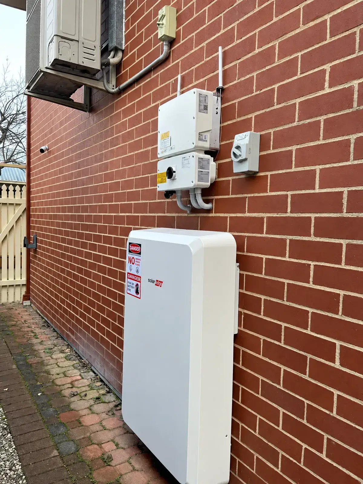 A tall white home battery unit stands against an exterior brick wall beneath electrical control boxes and conduit. An air-conditioning unit is mounted above to the left.